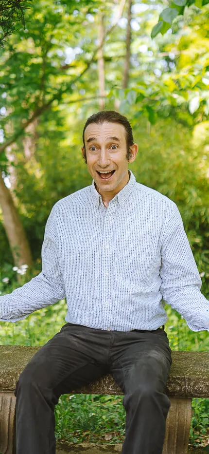 Abraham Burickson grinning wide-eyed in a pale checked shirt, surrounded by leafy green trees