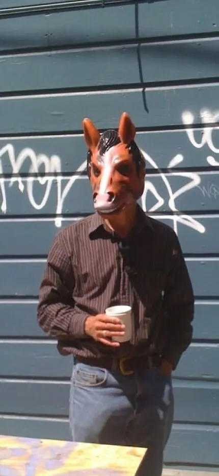 A man in a striped shirt and rubber horse mask standing before a graffitied blue wooden wall