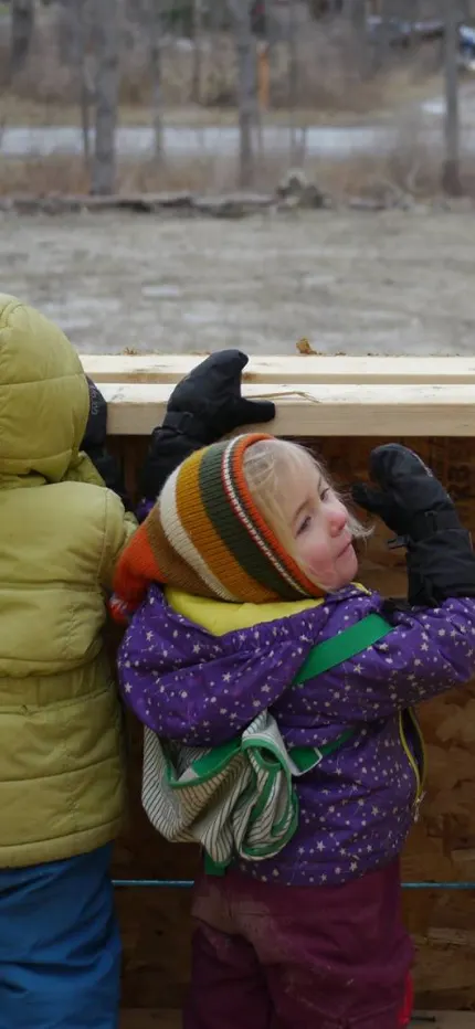 Two small children in winter coats looking out through the open wood framing of a house