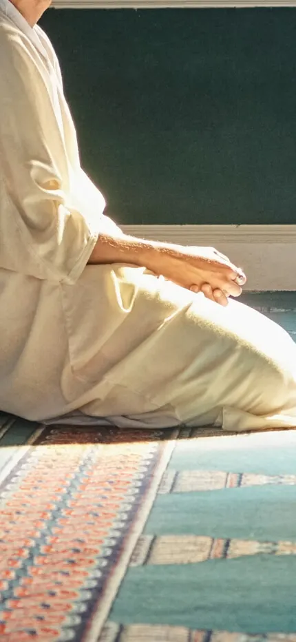 A man in a long white robe kneels on a patterned green prayer carpet in soft sunlight.