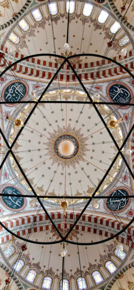 Looking straight up into an ornate mosque dome ringed with windows, beneath a star-shaped hanging chandelier frame.