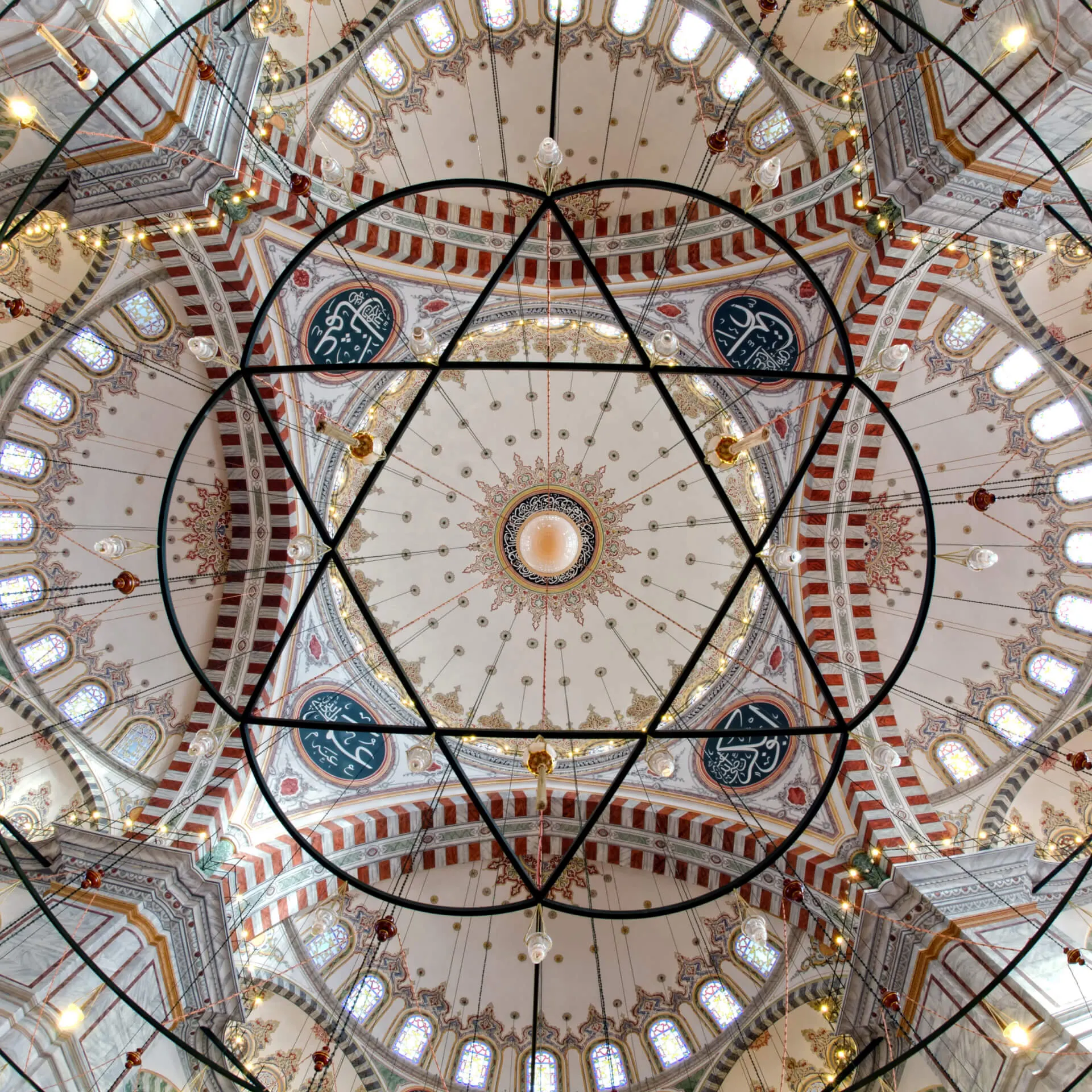Upward view of a painted mosque dome with calligraphy roundels, arched windows and a six-pointed chandelier frame.