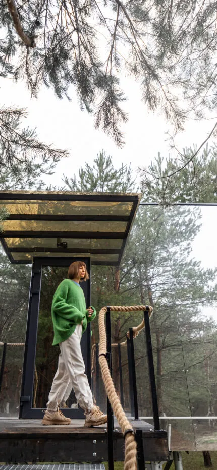 A woman in a green sweater stands on a small platform of a mirrored cabin among pine trees.