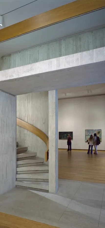 Gallery with a board-formed concrete spiral staircase, paintings on white walls and visitors looking at art.