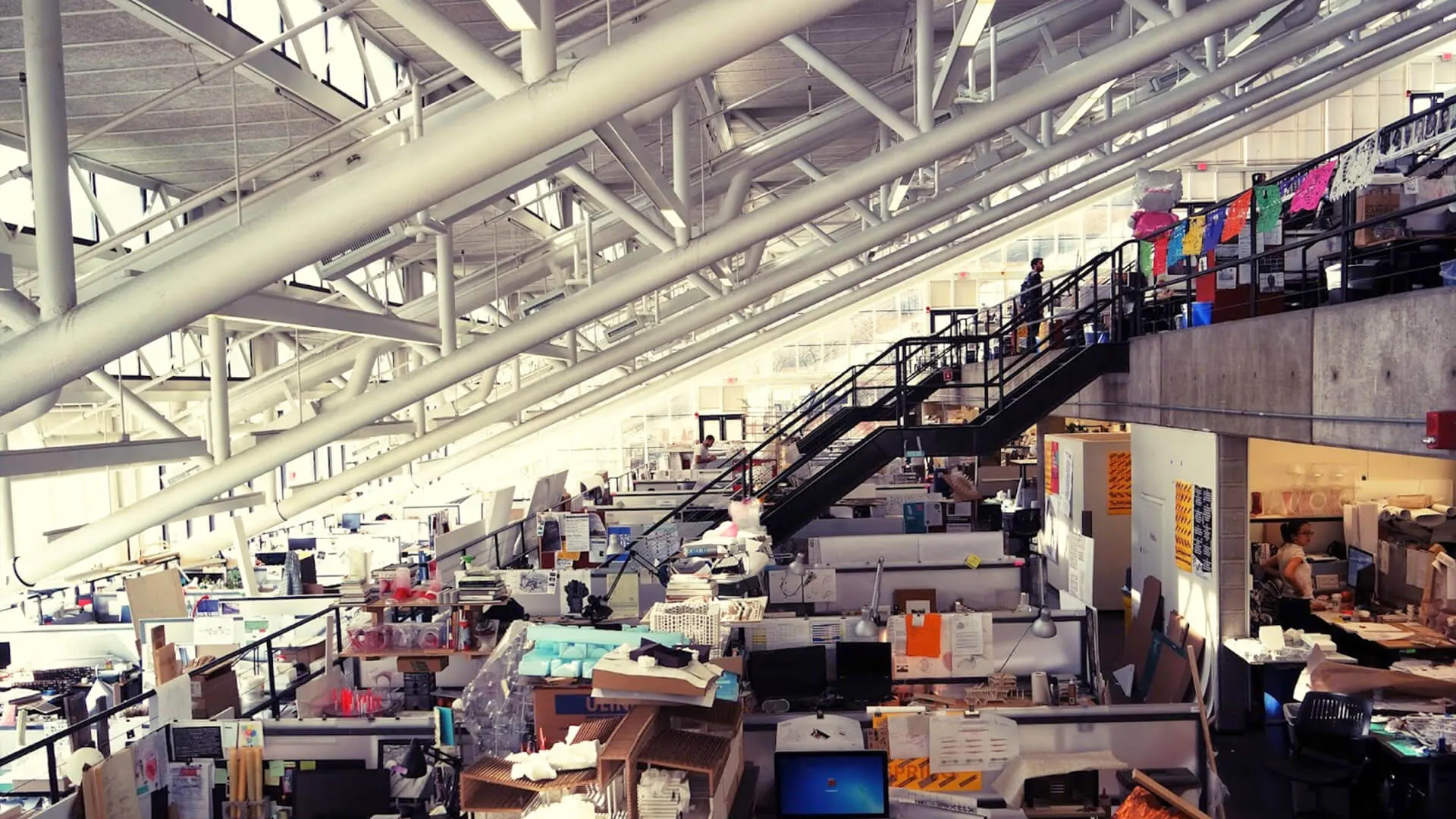 A vast, skylit design studio packed with desks, models and supplies beneath white steel trusses and stairs.