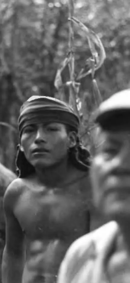 Black-and-white photo of a shirtless young man in a headband in the jungle, an older man in a cap blurred in front.