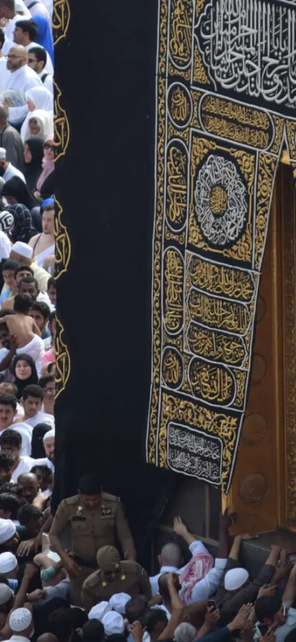 Dense crowd of pilgrims in white reaching toward the black, gold-embroidered cloth of the Kaaba door.