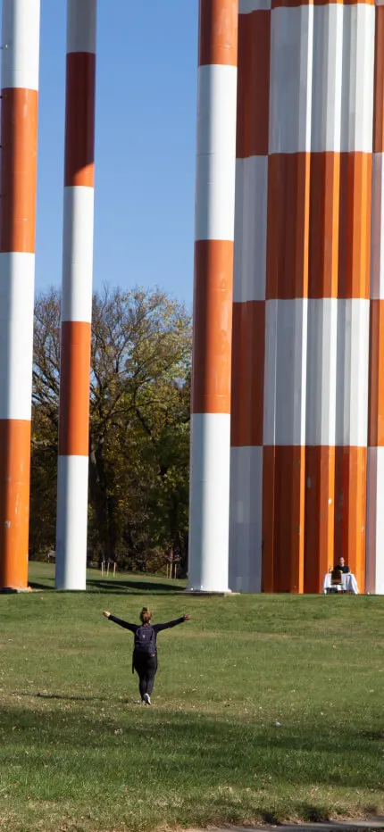 Lone person with outstretched arms on a lawn beneath towering orange-and-white striped columns.