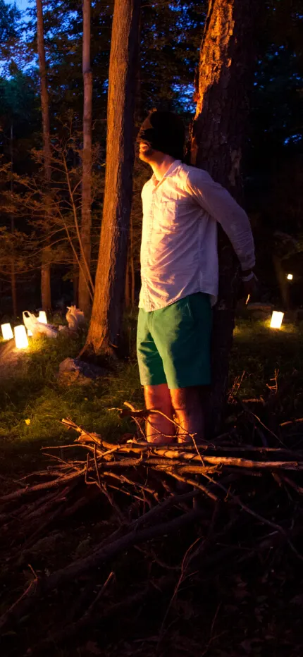Blindfolded man in a white shirt stands by a tree at a dusk forest clearing lined with glowing lanterns and a fire.