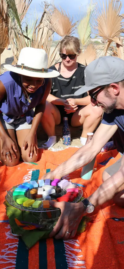 People in sun hats kneel around colorful blocks arranged on an orange cloth on a beach.