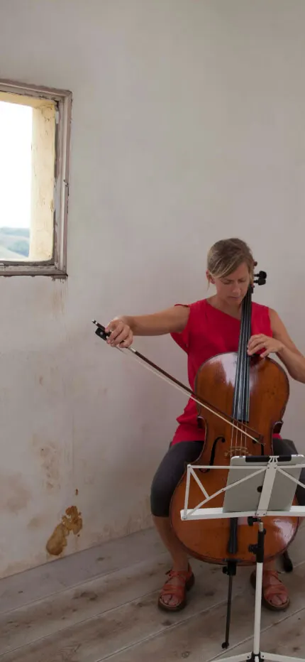 Woman in red playing cello before a music stand in a bare, weathered room with two windows.
