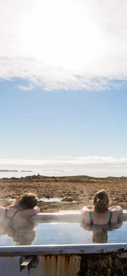 Two people soaking in a steaming outdoor hot tub facing sea, mountains and open sky