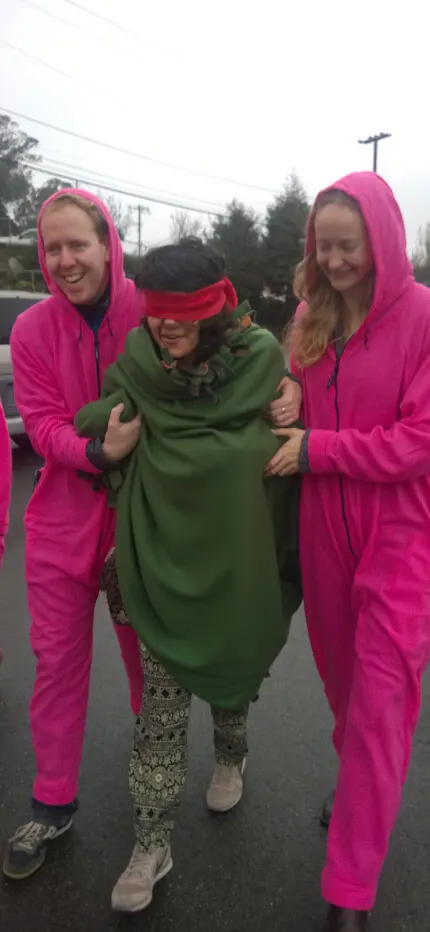 Smiling people in bright pink hoodies guide a blindfolded woman wrapped in a green shawl