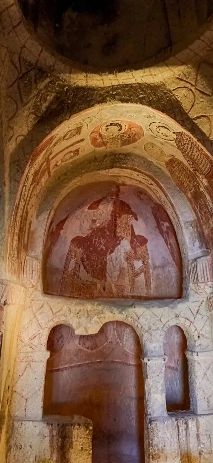 A dim rock-carved chapel alcove with faded red frescoes of a seated figure and haloed saints.