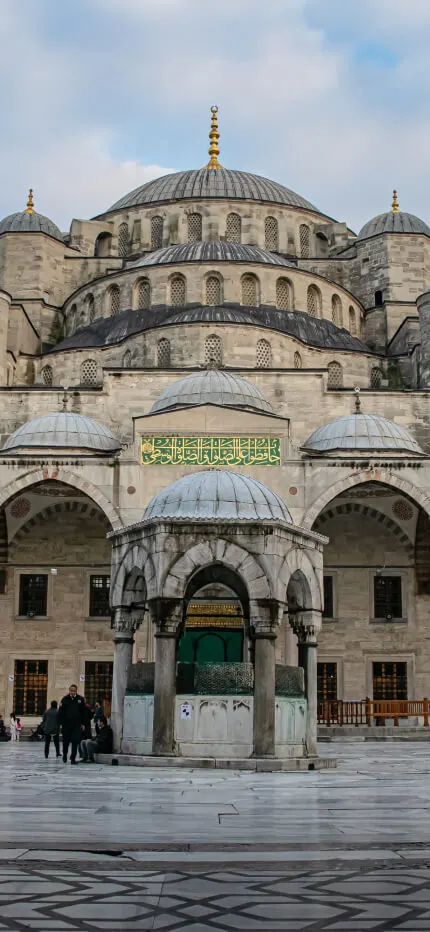 Mosque courtyard framed by dark columns, with cascading domes, slender minarets and a central domed fountain.