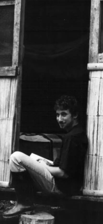 Black-and-white photo of a young man reading in the doorway of a bamboo-walled hut.